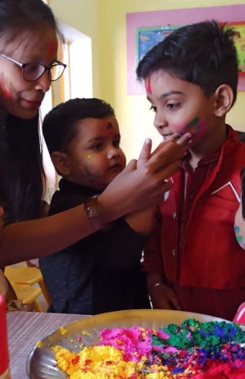 A teacher putting colour on a child's cheek during Holi, with a plate of gulal