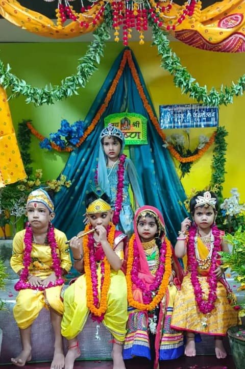 Children in Krishna and Radha costumes seated in front of the decorated Janmashtami stage