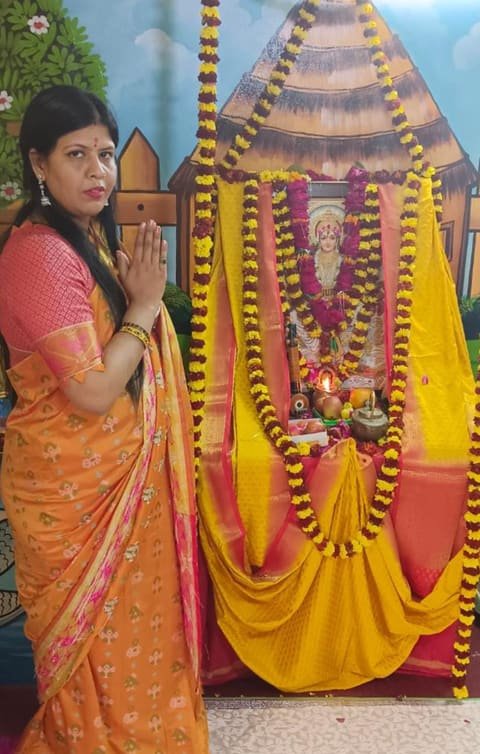 A teacher with folded hands beside a puja altar decorated with marigold garlands